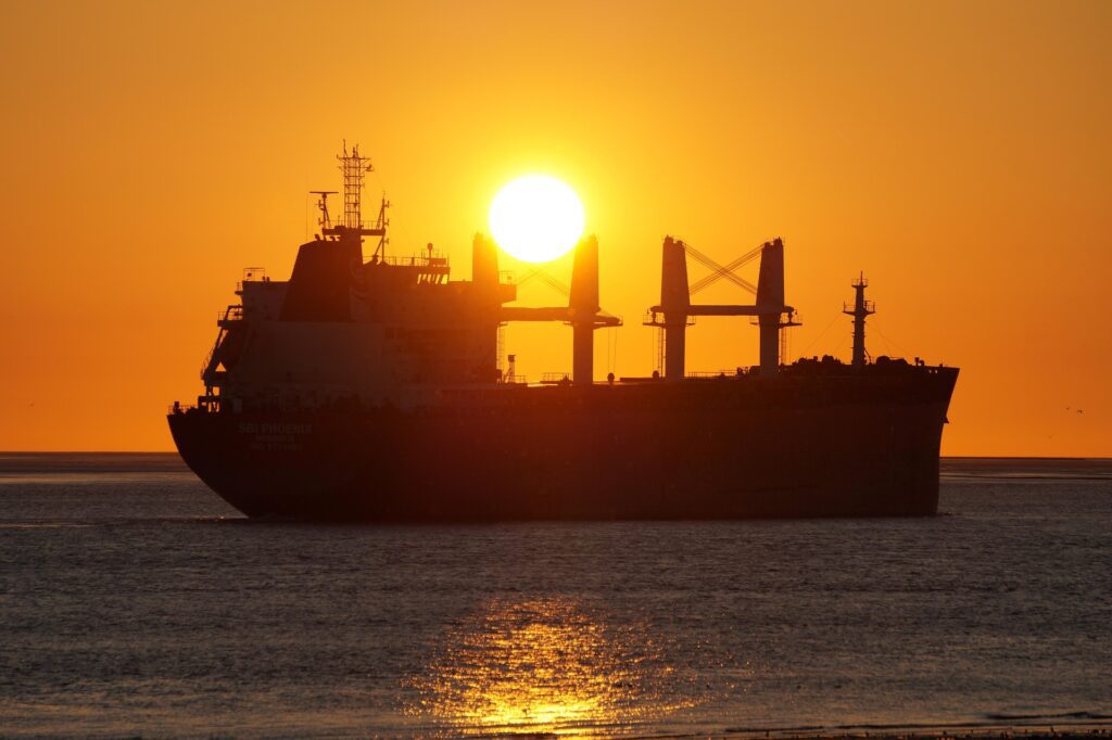 Large cargo ship silhouetted against a glowing orange sunset over calm ocean waters.