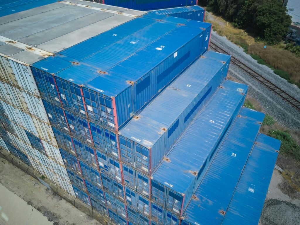 Aerial view of stacked blue shipping containers beside railroad tracks.