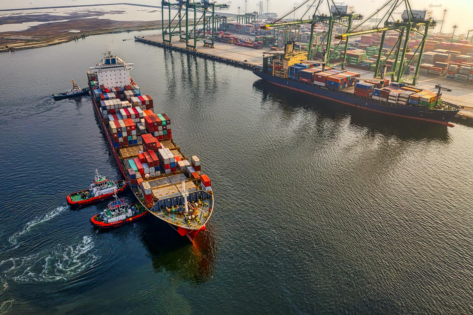 Aerial View of Container Ship at Industrial Port Container ship being assisted by tugboats near a busy shipping port with cranes and stacked containers.