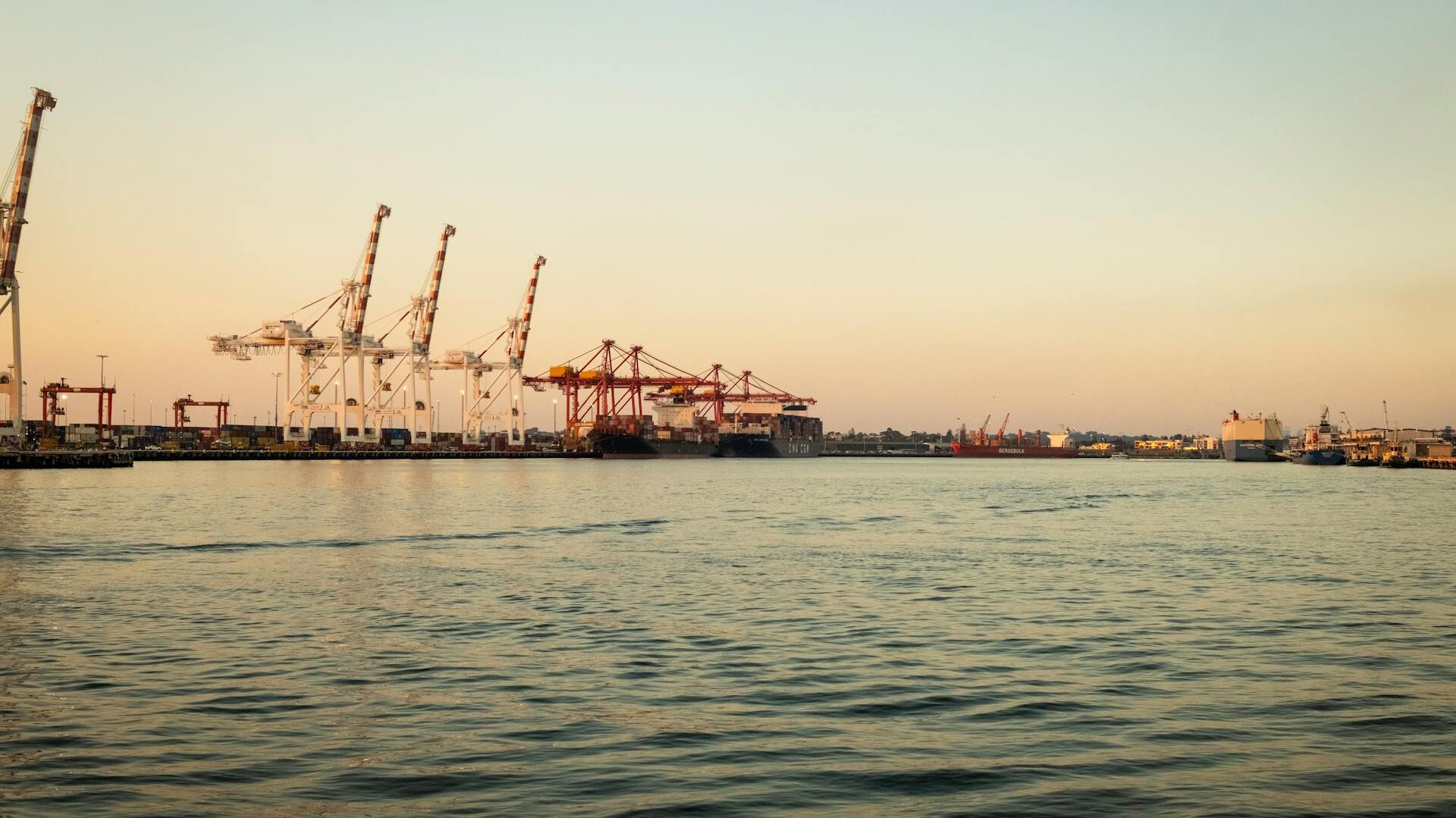 Container cranes and cargo ships at a busy industrial port during sunset.
