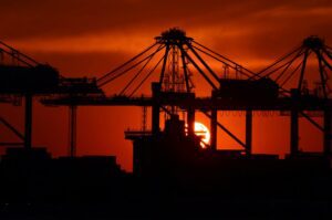 Silhouetted cranes and cargo ship at a port during a vivid red-orange sunset.