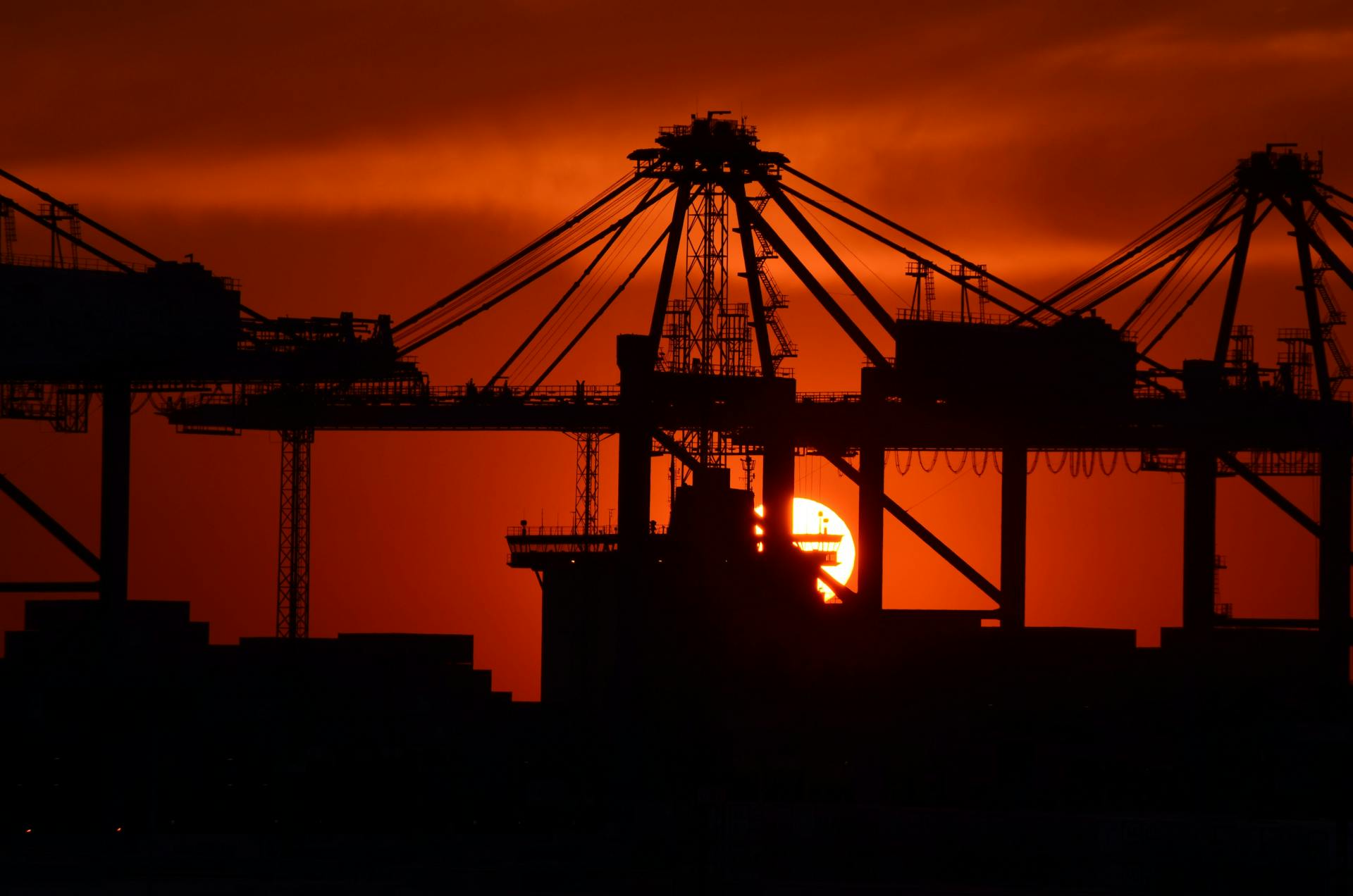 Silhouetted cranes and cargo ship at a port during a vivid red-orange sunset.